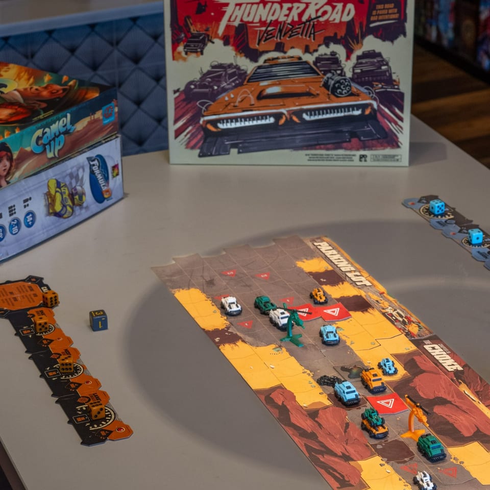 Board games "Thunder Road: Vendetta" and "Camel Up" with game pieces and dice on a table.